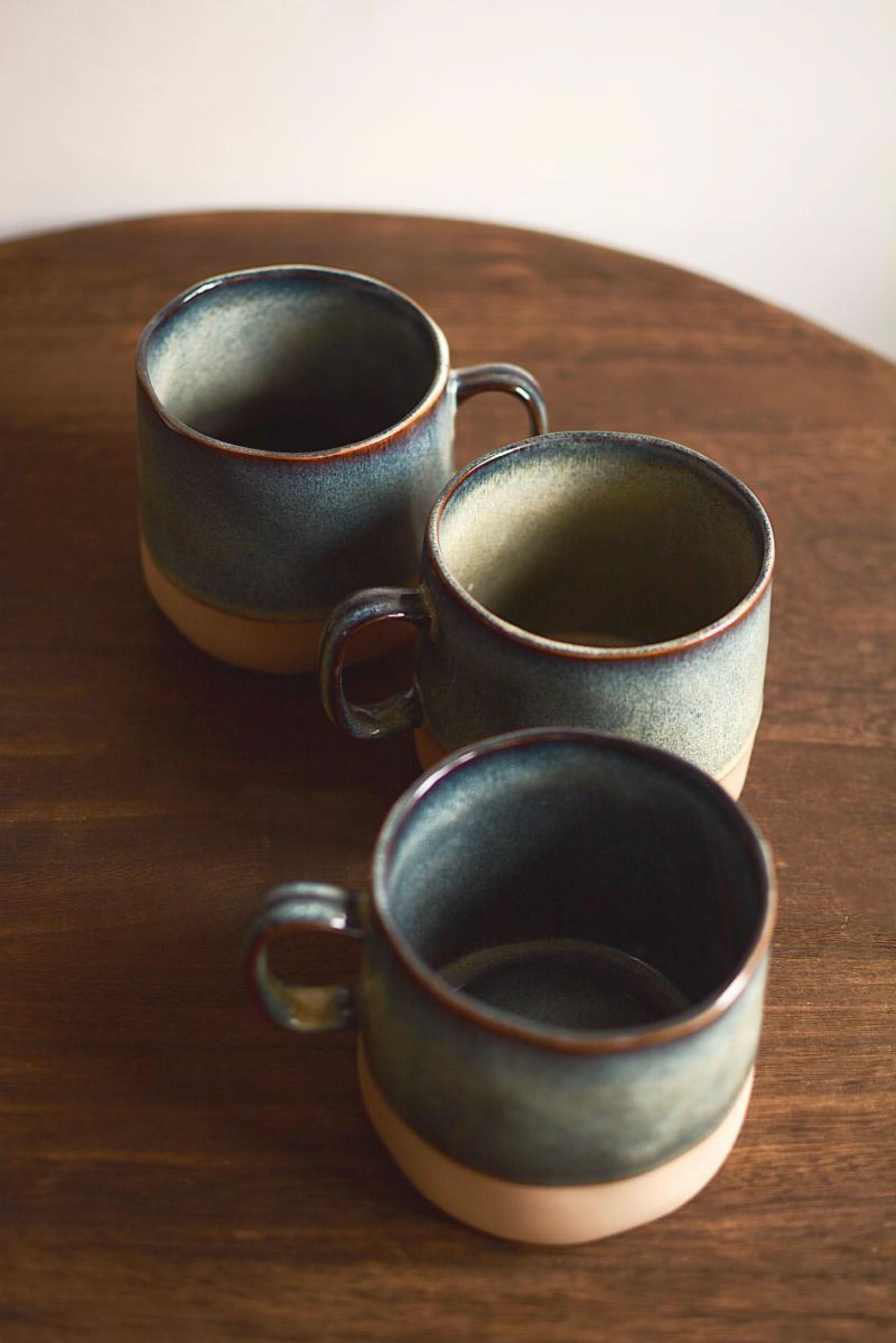 Set of 3 reactive glazed pottery mugs in blues and earth tones on a wooden surface.